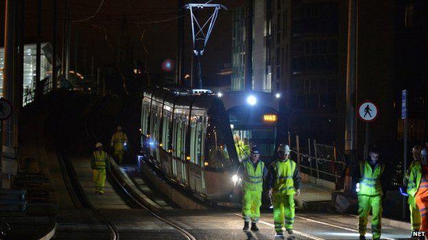 A tram crossing a bridge at night time with workers in high visibility clothing walking ahead of it.