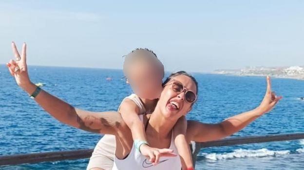 Mum in sunglasses has tongue out, smiling with daughter on her shoulders. Their arms are out making peace signs, there is a body of water behind them and blue sky. 