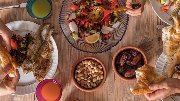 A variety of foods are laid out on a dining table, with a bowl of salad in the centre, a bowl of nuts and dates, and plates with meats to be shared.