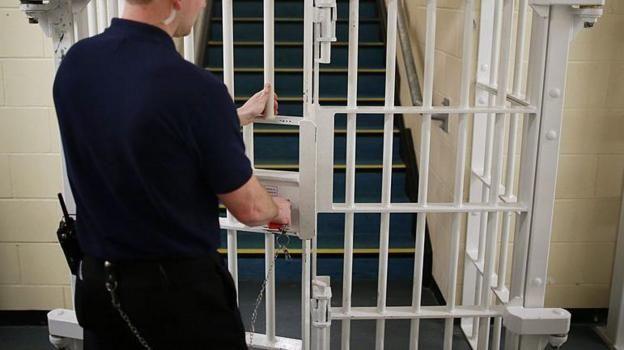 A man dressed in block with his hands on a white metal bar door. He has his hand next to a keyhole with a key in it.