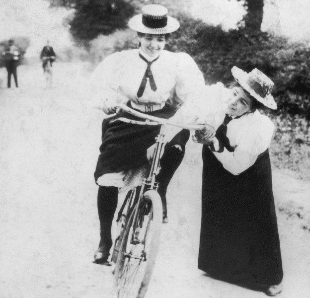 A young woman bicyclist is helped by a friend as she tries to maintain her balance, 1885