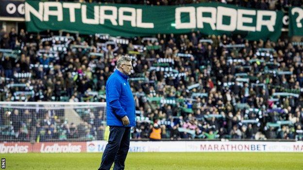 Hearts boss Craig Levein stand son the pitch as Hibs fans unveil a banner saying 'Natural Order'
