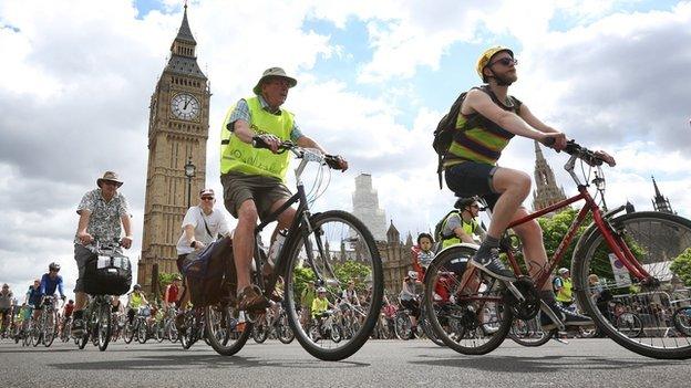 Lots of cyclists of all ages, cycling through London, Big Ben in the distance, behind them