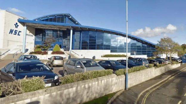 The exterior of the NSC, a large blue building with a curved roof with cars parked outside. There is also a pavement in front of a low white wall.