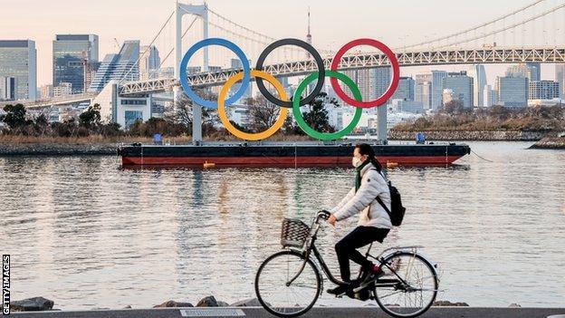 A woman wearing a facemask as a preventive measure against the spread of covid-19 rides past the Olympic logo