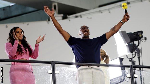 Ian Wright celebrates with his hands in the air and a BBC Sport microphone as Alex Scott claps