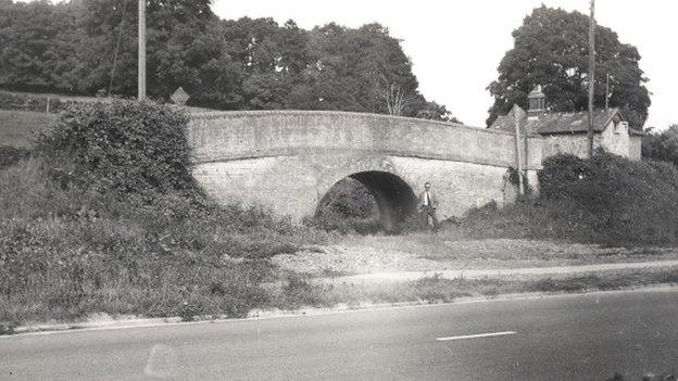 View shows a man standing by the bridge crossing a disused canal, the bridge is alongside a road which crosses where the canal would have been, taken in the 1950s.