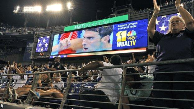 Fans cheers as video of United States" Michael Phelps celebrating winning gold in the men"s 200-meter individual medley during the swimming competitions at the Summer Olympics is re-played on the large television screen, during the second half of an NFL preseason football game between the Baltimore Ravens and the Carolina Panthers,