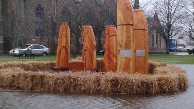 A carved wooden nativity scene is surrounded by water in Ballater