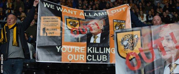 Hull City fans hold banners protesting against Assem Allam. The first banner reads We Want Allams Out, You're Killing Our Club, and Bring Back Concessions. The other has the word Out in front of a picture of Allam