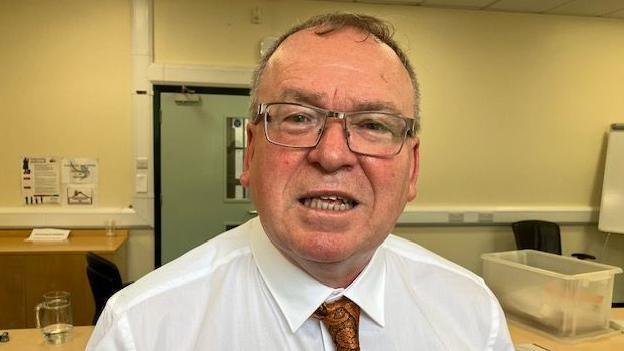 Mae Clive yn edrych ar y camera. Mae'n gwisgo crys gwyn a thei lliw efydd gyda phatrwm paisley arno. Mae gan Clive sbectol a gwallt brown byr. Gellir gweld desg y tu ôl iddo.