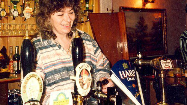 Deborah Steel working behind a bar, pouring a pint. She is smiling as she does it and wears a stripy blue and pink patterned jumper. The taps in front of her read Abbot Ale, Harp Larger and Greene King.