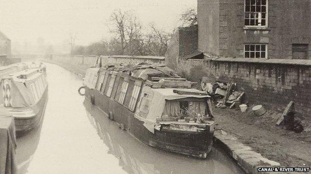 Photograph of 'Poo-Koo', an early residential conversion of a wooden narrowboat, moored at Banbury on the Grand Union Canal. Taken showing the bow of the boat with another narrowboat opposite it on the left of the photograph and a building on the right. 1957-1962