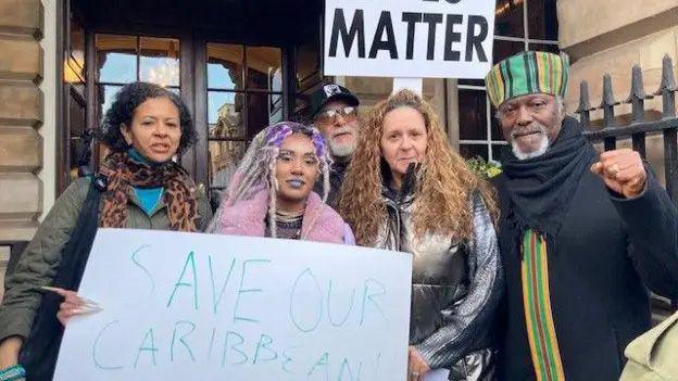 Three women and two men stand outside the door of Liverpool Town Hall holding a sign saying Black Lives Matter and Save Our Caribbean Centre.