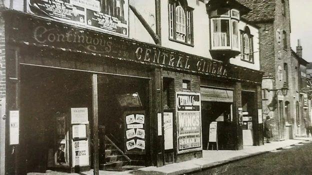 Black and white photo of two storeys of an art-deco cinema in a building that is clearly older than the cinema which opened in 1930. The ground floor is dark in colour with light-coloured letters spelling Continuous CENTRAL CINEMA above the doorways. There are stairs going upwards on the left side of the entrance and several advertising posters are visible.