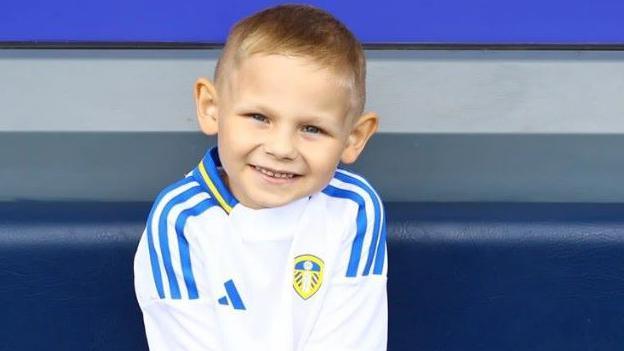 Bob, six, sat on a blue bench, wearing a white and blue football kit 