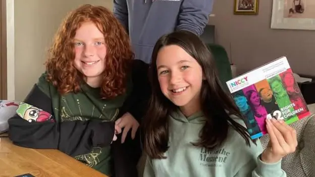 Two girls sit at a table - one has curly red hair, the other has straight dark hair. The girl on the right is holding a pamphlet.
