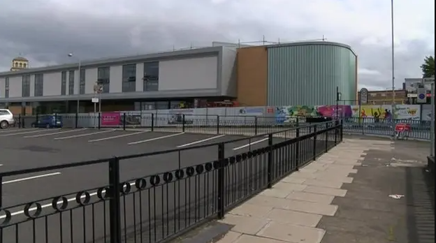 The youth club building is curved at one end with a light blue-green facade. The other part of the building to the left is grey with large windows. There is a car park in front of the building, with railings in the foreground of the shot.