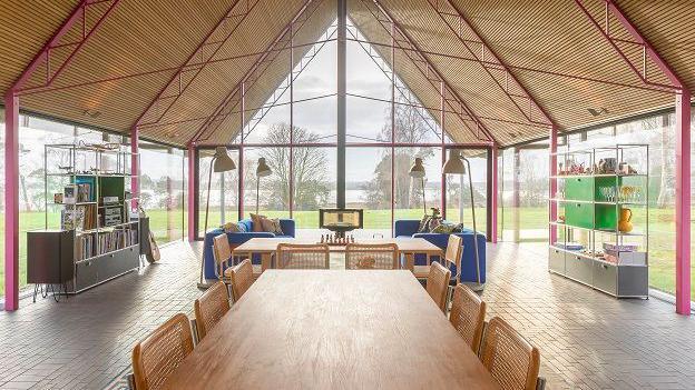 Large wide glass-walled room with wooden triangular ceiling. There is a large wooden fable with wooden chairs in the foreground and a further table beyond. There are storage units to the right and left.