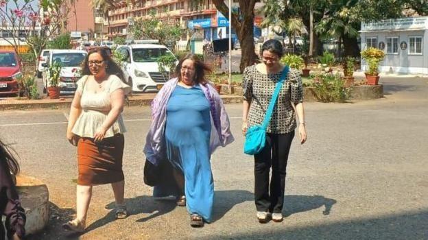 Three women walking across a road in Goa, all have long brown hair, woman on left is wearing cream blouse and brown skirt with sandles, woman in middle is wearing blue dress with purple scarf, woman on right is in checkered black and white top with black trousers, a bright blue crossbody bag and sandles

There are plants and vehicles visible in the background