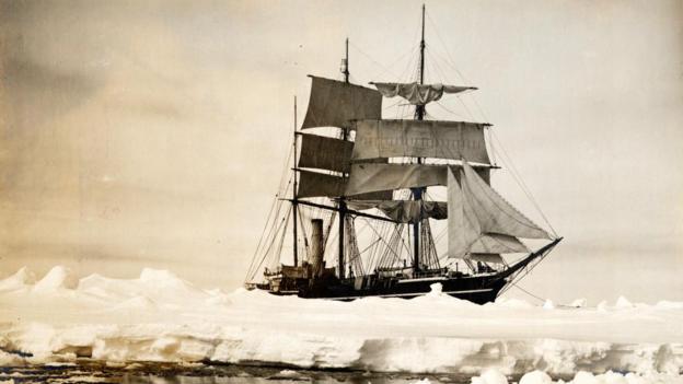 Black and white image of the Terra Nova ship sailing in Antarctica. The ship is wooden, it's sails are open. It is moving across ice-covered ocean.