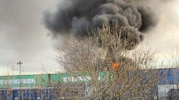 Huge plume of dark smoke seen above storage containers. There is the orange glow of some flames visible lower down. The image is partially obscured by a large bush with no leaves on it. 