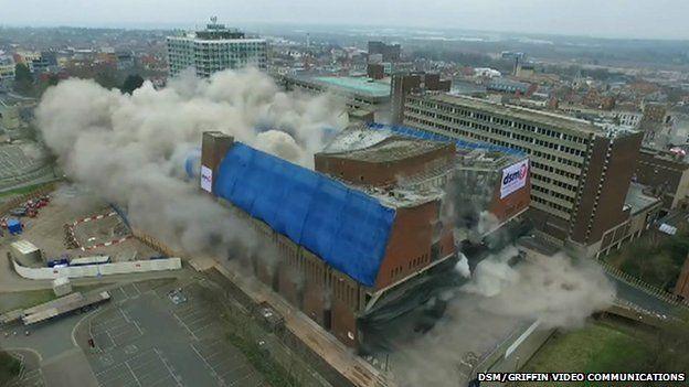 Northampton's Greyfriars Bus Station being demolished. The building has blue covering on one side of its roof. Grey clouds of debris are rising from the structure as detonators go off