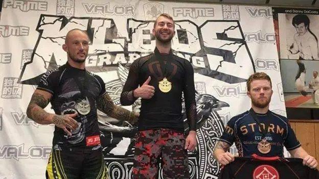 Will Stone pictured at a MMA competition. He stands in between two men and poses with a medal around his neck.