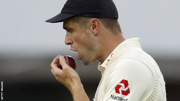 Side on shot of Chris Woakes in England Test kit and cap, holding the ball up in front of his mouth