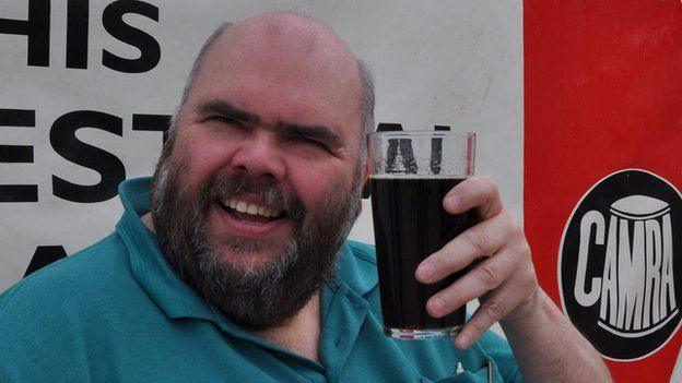 A man holds a pint up as he smiles at the camera. He is bald and has a thick beard. He is wearing a blue polo top. There is a "Camra" logo in the background.