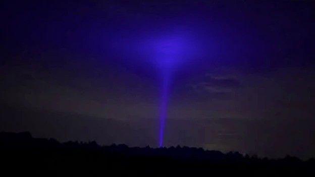 A beam of blue light shoots upwards and fills a dark cloudy night sky above some trees