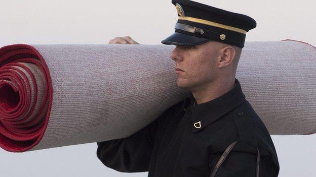 A red carpet is carried by a military officer