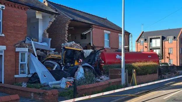 A tractor pulling a red tanker crashed into two terraced houses. The front of the houses is totally destroyed.