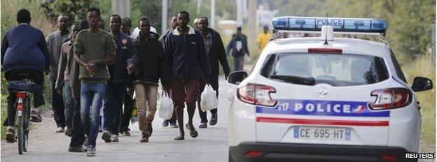 French police patrol as migrants walk on a street near the makeshift camp called "The Jungle" in Calais