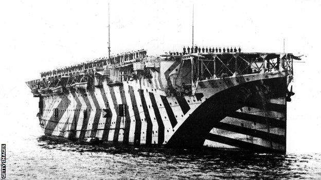 November 1918: Airmen and seamen cheering King George V from the aircraft carrier "Argus" on his visit to the Fleet at Rosyth, on the Firth of Forth. The carrier is painted in "dazzle" camouflage, designed to confuse enemy ships - not so much to hide vessels, instead the camouflage was intended to make it difficult to pinpoint the direction in which a ship was travelling.