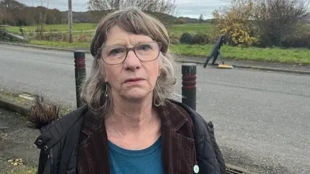 A woman wearing glasses with greying brown hair is wearing a jacket and a blue top. She is standing next to a road with fields in the background.