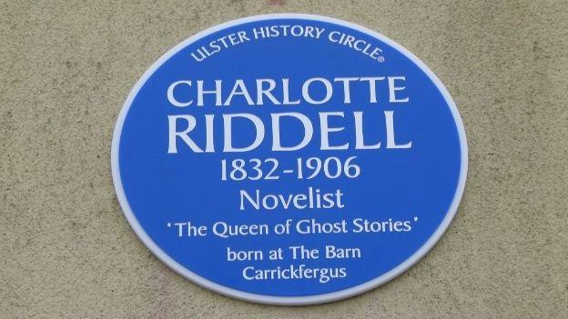 A blue Ulster History Circle plaque which says "Charlotte Riddell 1832 - 1906 Novelist 'The Queen of Ghost Stories' born at The Barn Carrickfergus". 