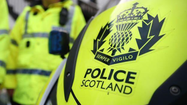 Wording which says 'POLICE SCOTLAND' in bold letters on a yellow police motorbike, with an officer in the background.