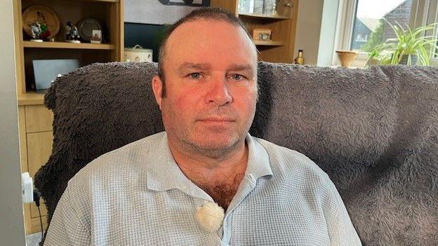 Lee sitting on a sofa in a living room with a window behind him