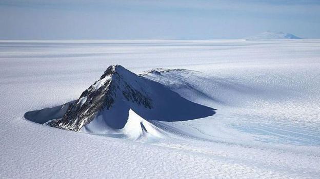 A section of the West Antarctic Ice Sheet with mountain peaks poking out of the pristine, flat, white icy surface. The sky above is light blue with light clouds.