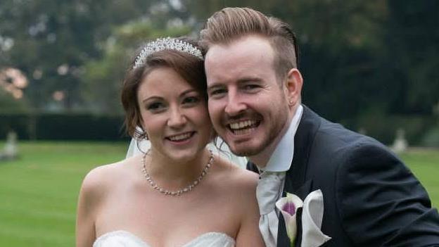Jim and Stephanie Rugg on their wedding day. Jim has fair hair, is laughing and is wearing a black suit jacket with a white shirt, tie and a flower on his chest pocket. Stephanie is wearing a white wedding dress with a necklace and tiara. she is smiling and has brown hair.