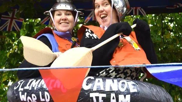 Two people pose for a photo in a carnival float designed to look like an inflatable canoe. They are wearing metal colanders for helmets and have cardboard paddles. They are surrounded by bunting.