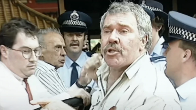 Still from the video, showing Jack Karlson, wearing a beige shirt, surrounded by police and men in shirts trying to hussle him along as he speaks to the camera.