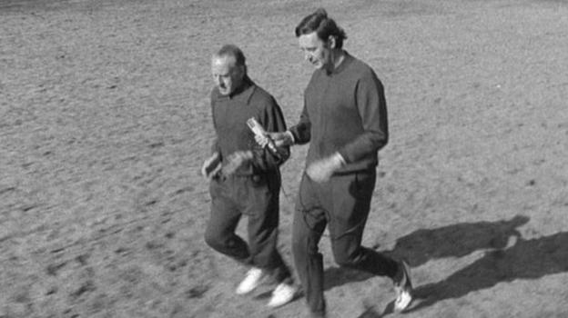 Two men wearing tracksuits jog across the grass in the park.