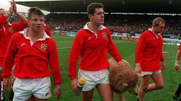 Gavin Hastings, carrying a Lions mascot, and flanked by Rob Andrew (left) and Dean Richards (right), walks off the pitch after the Lions' third Test defeat against New Zealand in 1993