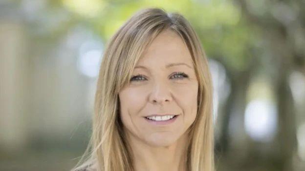 Close up of woman's face, she has blonde hair and is smiling while stood outside with blurred leaves in the background.