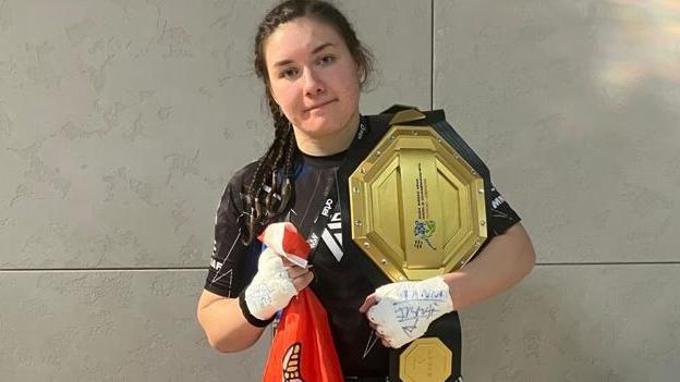 Alanna Pritchard looking at the camera. She is wearing a sports top and is carrying a large gold belt over her right shoulder. He hands are taped up in white tape and she is has a red Manx flag in her left hand.