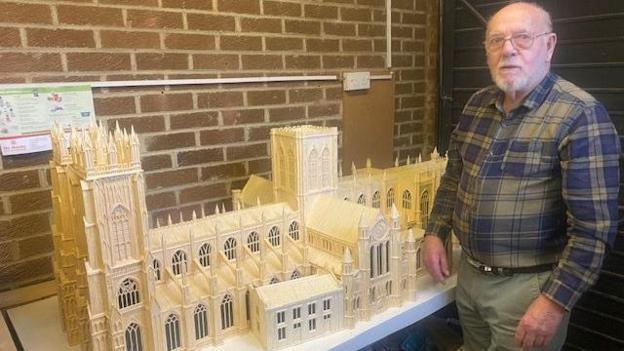 An older man wearing a checked shirt stands next to a model of York Minster which is made from matchsticks.