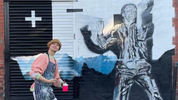 Artist Jack Brett poses near his mural wearing a painting apron and holding a pot and brushes.