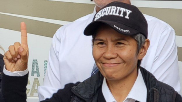 Romana Didulo wearing black cap with word "security"in white, holding up index finger.\